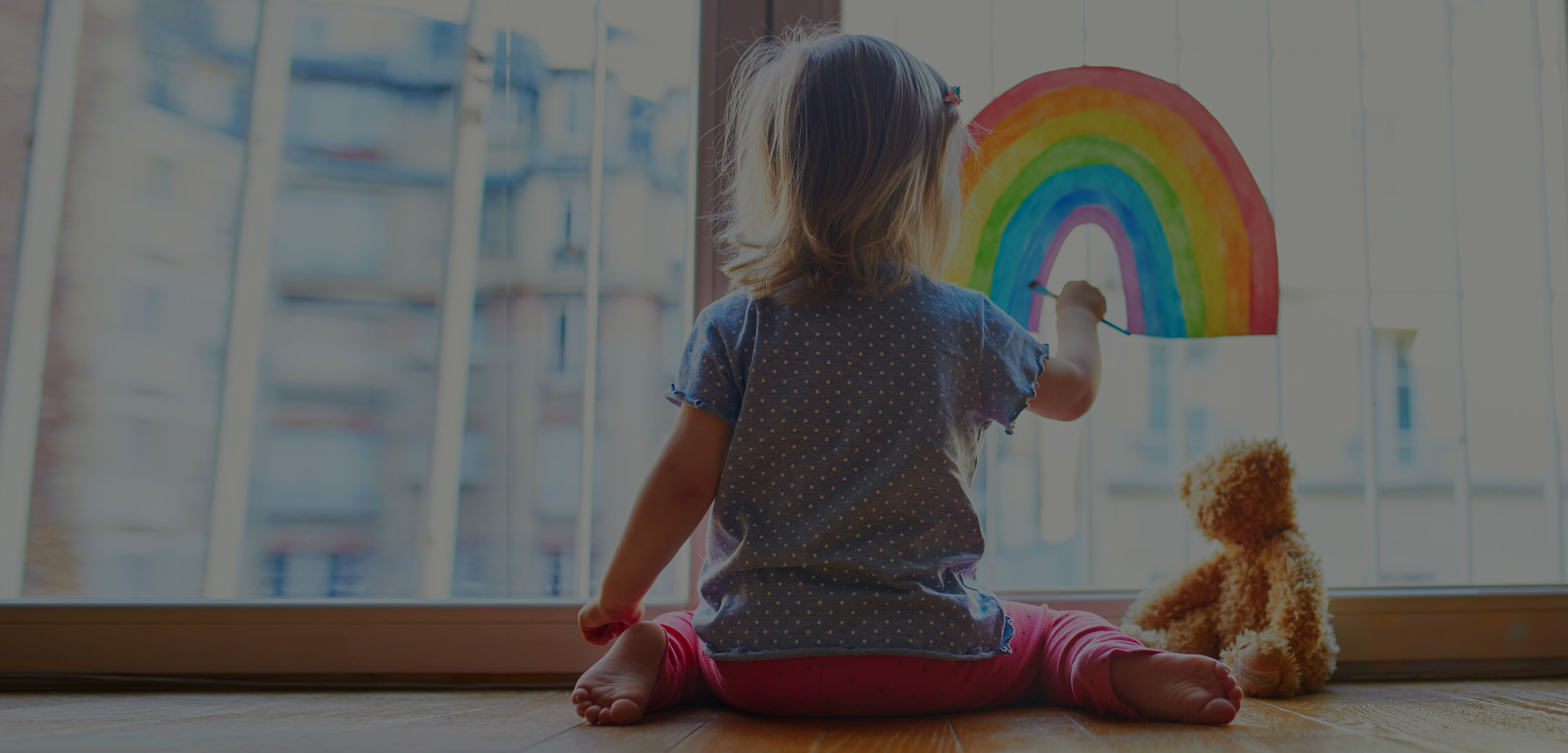 Young nursery school child, with her back to us, draws a rather impressive rainbow with paints on the glass window as her toy bear looks on