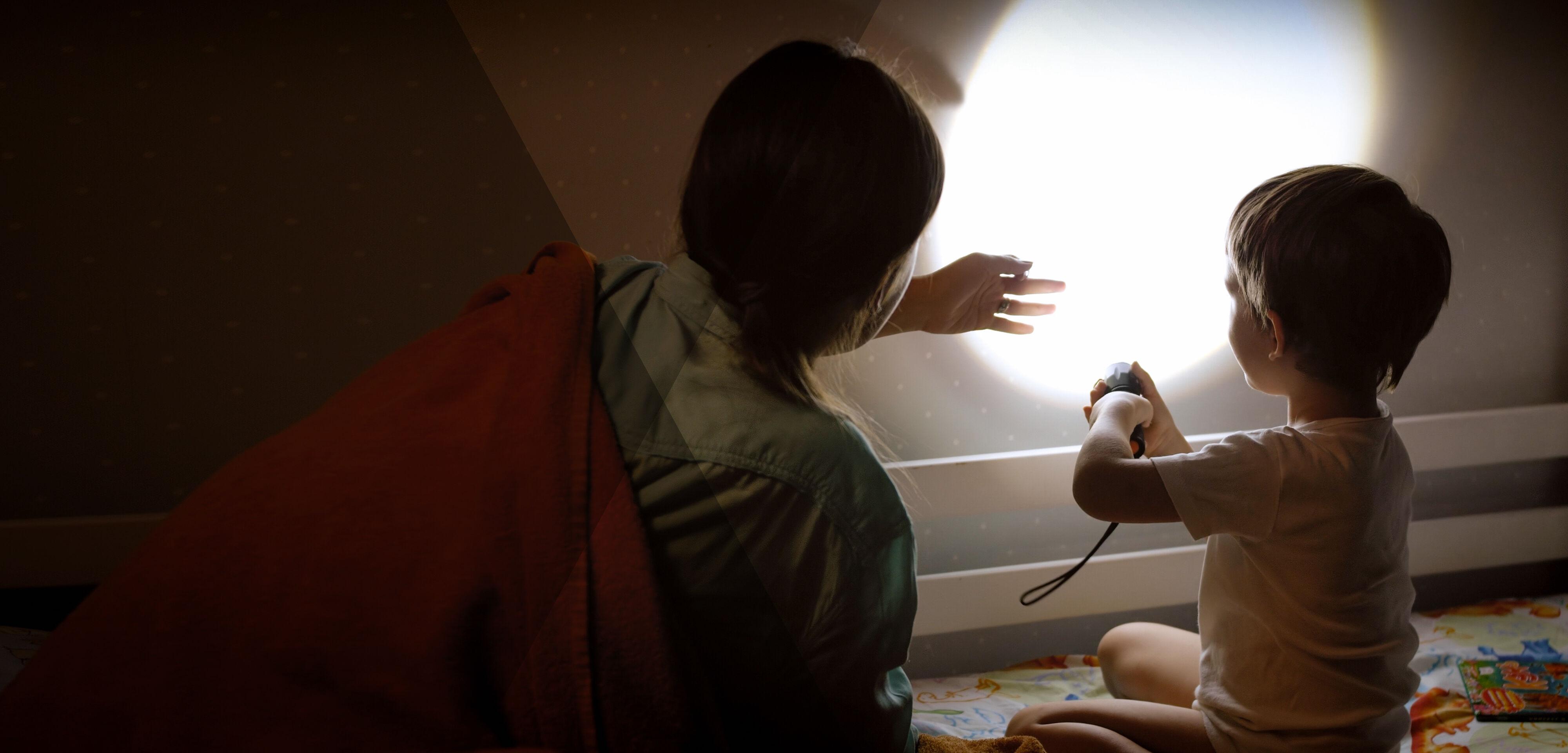 Young child sits on the floor with his mother, shining a bright torch on to the wall, casting long shadows within the room