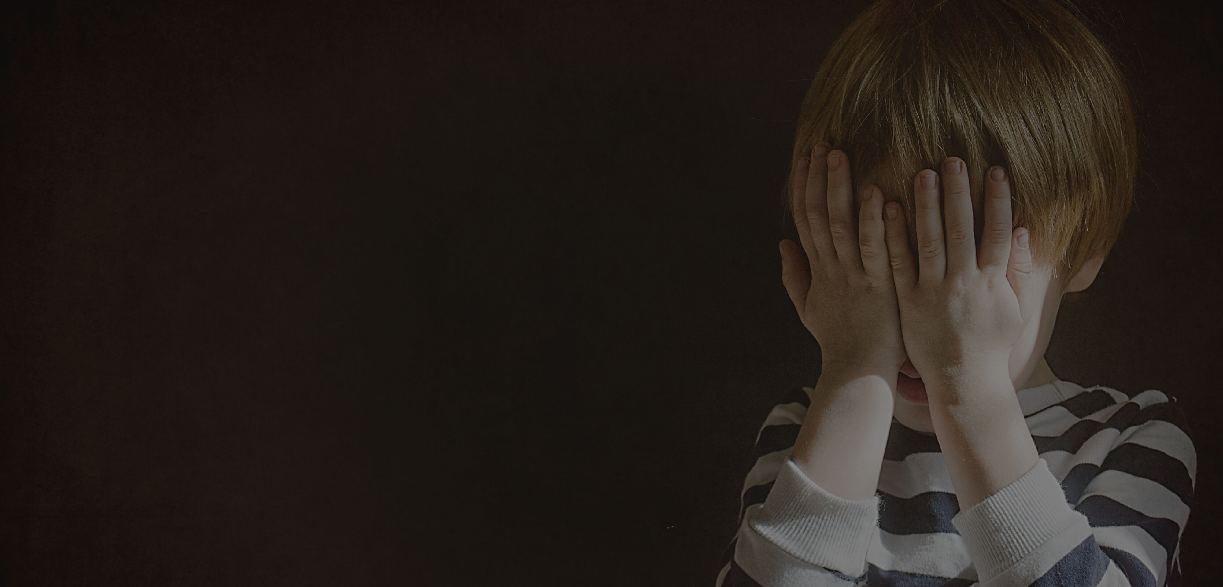 Small sad male child in striped jumper covers his face with two hands in a darkened room