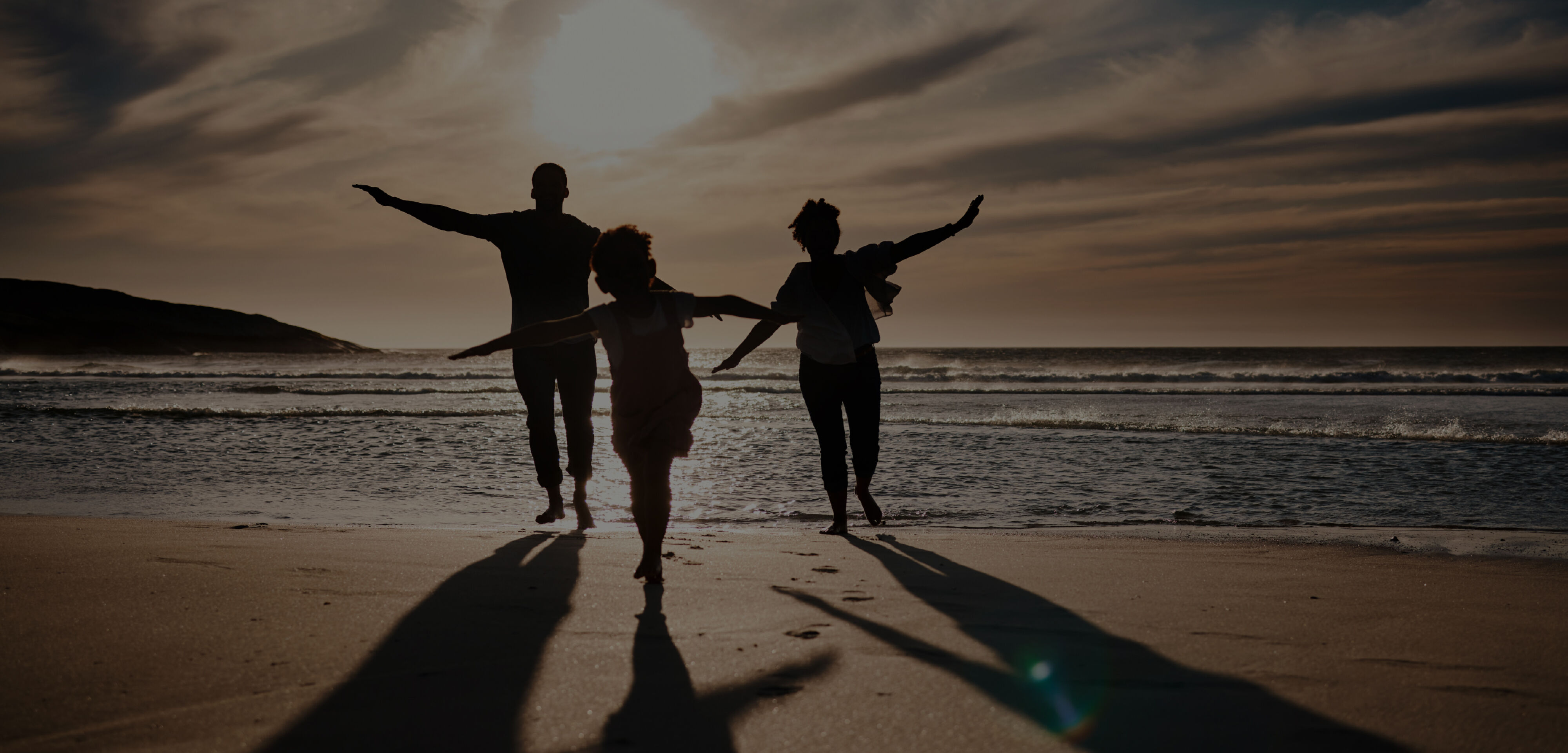 Three Silhouettes stand on a beach with the sun casting their long shadows on to the sand