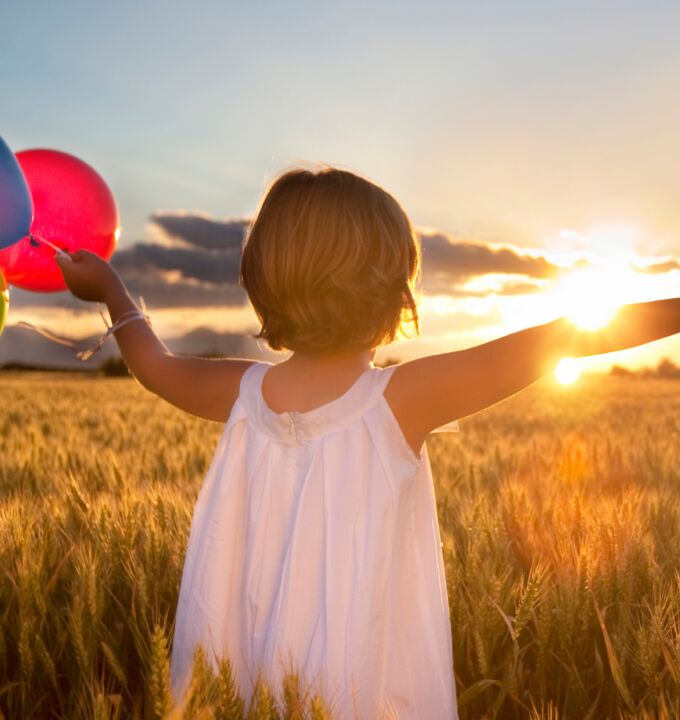 Young Child stands in a wheat field holding multi coloured balloons