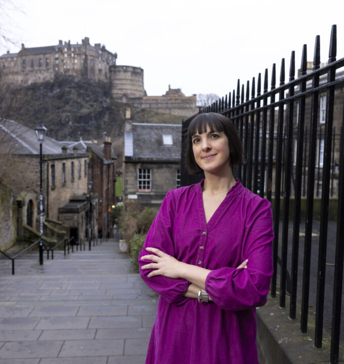 Zoe, in her magenta dress stands resting on black railings with Edinburgh Castle in the background