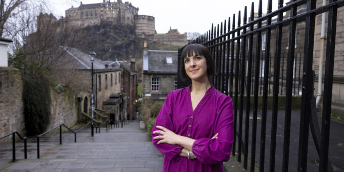 Zoe, in her magenta dress stands resting on black railings with Edinburgh Castle in the background